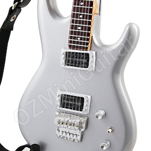 Puede incluir: Una guitarra el&eacute;ctrica plateada con una correa negra y un soporte. La guitarra tiene dos pastillas humbucker y un puente tremolo.