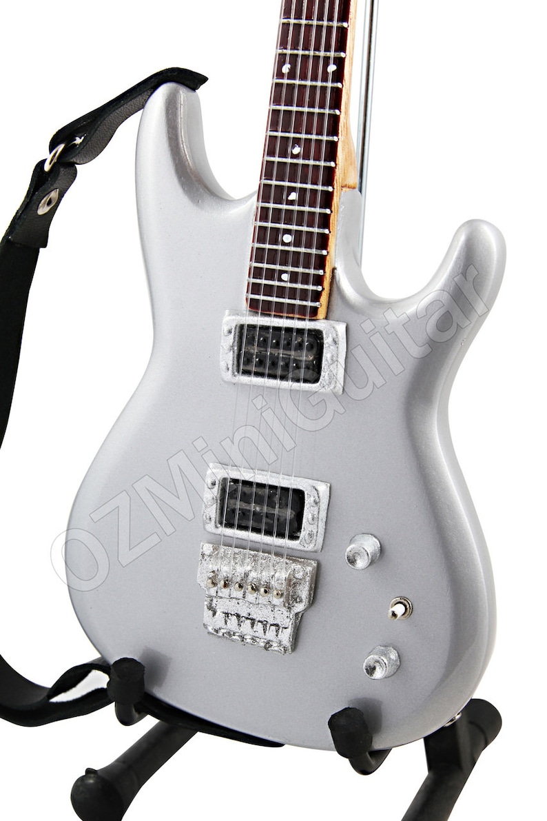 Puede incluir: Una guitarra el&eacute;ctrica plateada con una correa negra y un soporte. La guitarra tiene dos pastillas humbucker y un puente tremolo.