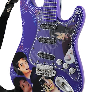 Puede incluir: Una guitarra eléctrica morada con una golpeador blanco y una correa. La guitarra está decorada con imágenes de Michael Jackson y tiene herrajes plateados.