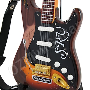 Op de afbeelding: Een bruine en zwarte elektrische gitaar met een gouden slagplaat en een witte band. De gitaar heeft een zwarte slagplaat met de tekst "S.R.V. Custom" erop.