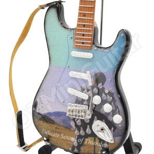 May include: A miniature electric guitar with a light blue and black finish, featuring a landscape design and the text "Delicate Sound of Thunder." It has white pickups, a silver bridge, and a brown strap. The guitar is displayed on a black stand.
