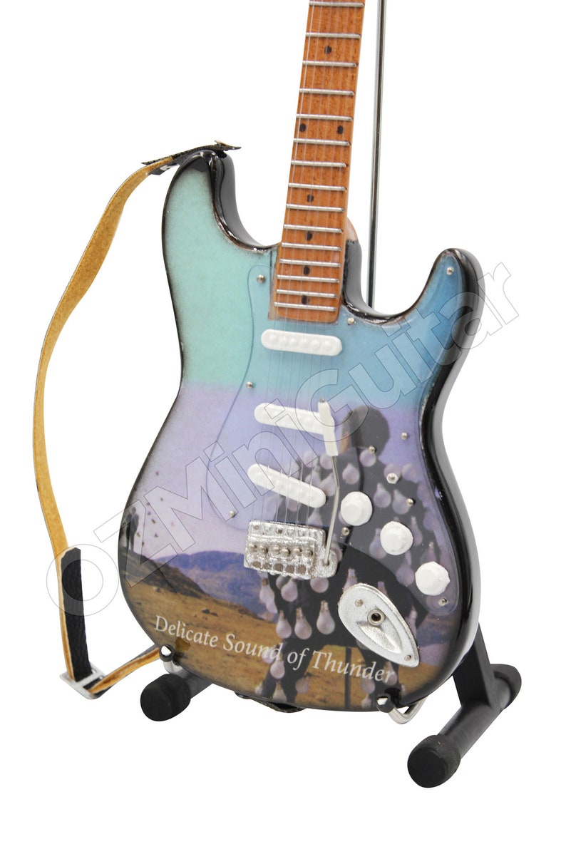 May include: A miniature electric guitar with a light blue and black finish, featuring a landscape design and the text "Delicate Sound of Thunder." It has white pickups, a silver bridge, and a brown strap. The guitar is displayed on a black stand.