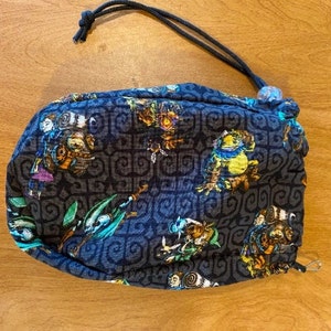 May include: A black drawstring bag with a dark gray background and a repeating pattern of stylized figures. The figures are in shades of blue, green, yellow, and orange. The bag has a black drawstring and a small, round, purple bead at the top.