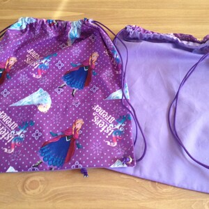 May include: Two purple drawstring bags. One bag features a pattern of a princess, snowflakes, and the text "Sisters forever" in white. The other bag is a solid light purple color. Both bags have purple drawstrings.