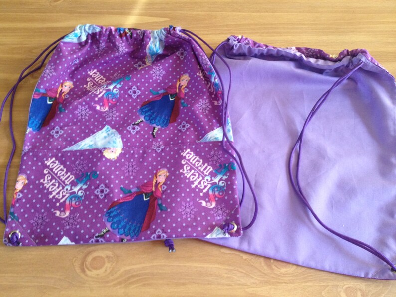 May include: Two purple drawstring bags. One bag features a pattern of a princess, snowflakes, and the text "Sisters forever" in white. The other bag is a solid light purple color. Both bags have purple drawstrings.