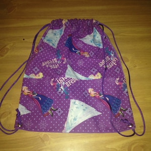 May include: Purple drawstring backpack featuring a repeating pattern of a cartoon character in a blue dress, snowflakes, and the text "Sisters Forever". The bag has a purple drawstring closure.