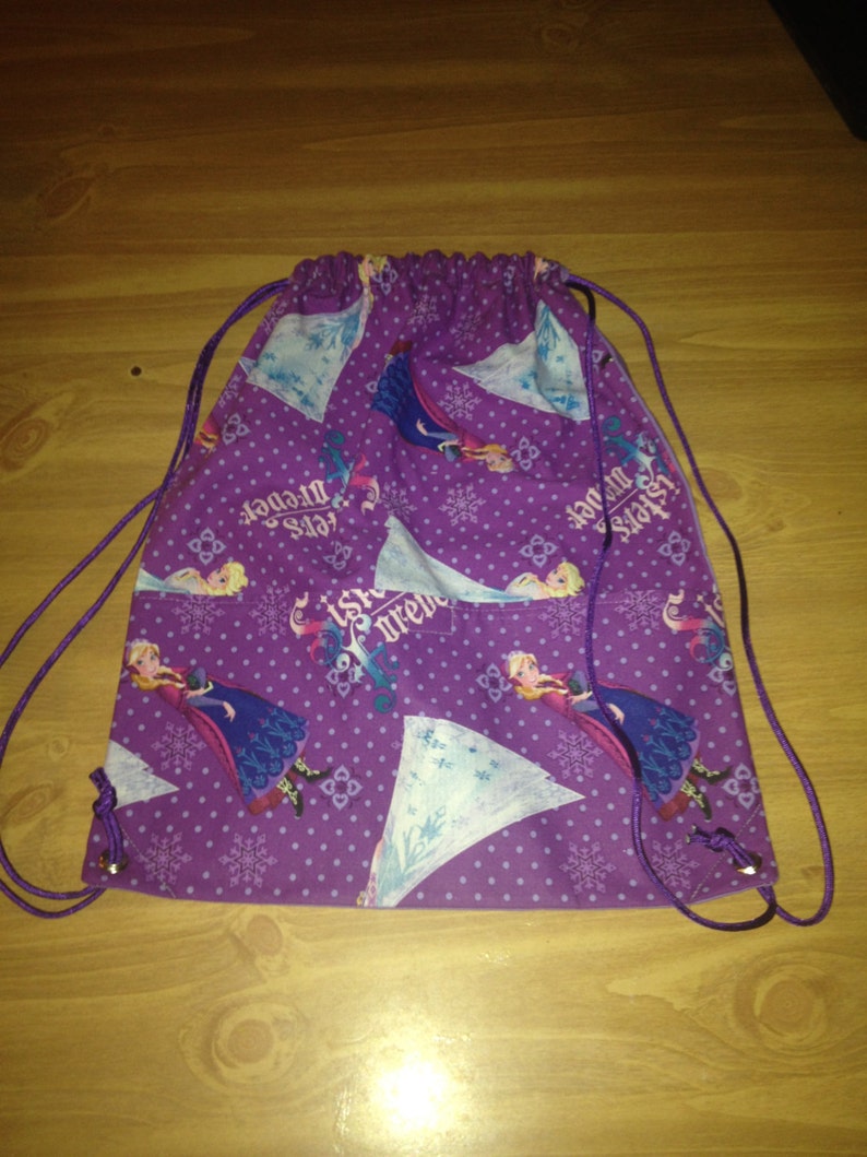 May include: Purple drawstring backpack featuring a repeating pattern of a cartoon character in a blue dress, snowflakes, and the text "Sisters Forever". The bag has a purple drawstring closure.