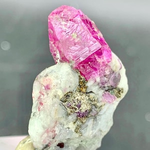 May include: A striking pink ruby crystal, set within a white and grey matrix. The ruby's crystalline structure is visible, highlighting its natural form. The base of the specimen is a light tan colour, with a grey background.