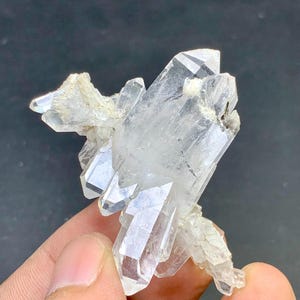 Puede incluir: Un grupo de cristales de cuarzo transparente con terminaciones puntiagudas. Los cristales son translúcidos y tienen un aspecto natural y en bruto. La formación de cuarzo se encuentra sobre un fondo oscuro, resaltando su estructura cristalina.