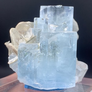 Puede incluir: Un grupo de cristales de aguamarina azul claro con un aspecto transparente y vítreo. Los cristales están dispuestos en forma de pirámide y se colocan sobre una base azul oscuro.