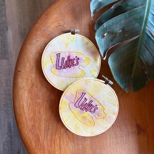 May include: Two yellow and pink tie-dye embroidery hoops with the text "Luke's" in purple embroidery.