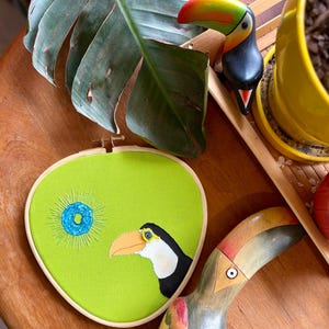 May include: A green fabric embroidery hoop with a toucan and a blue flower design. The hoop is on a wooden surface with a wooden toucan and a yellow planter with a green plant.