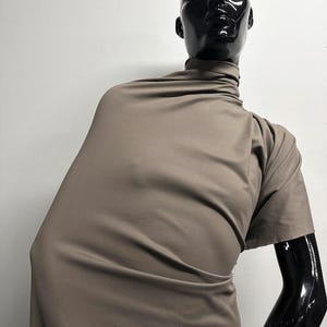 May include: A taupe-colored fabric draped over a black mannequin. The fabric is wrapped around the mannequin's neck and shoulders, forming a draped effect. The mannequin has a glossy black finish, and the background is a plain white wall.