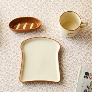 Puede incluir: Un juego de vajilla de cerámica con temática de pan, que incluye un plato con forma de tostada, un plato ovalado pequeño y una taza. El plato y la taza tienen ribetes marrones. El plato tiene la palabra "bread" impresa.