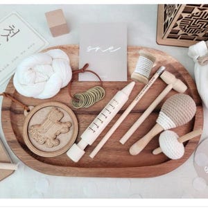 May include: A wooden tray with a variety of items, including a white crocheted rope, a wooden toy, a wooden syringe, a wooden hammer, a wooden rattle, a spool of thread, and a pile of gold coins. The tray is on a white surface. The text "매쓰" is visible on a piece of paper.