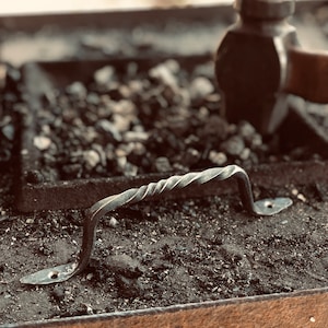 May include: Close-up of a hand-forged metal handle with a twisted design. The handle is dark gray and has a curved shape with mounting plates on each end. The background shows a blacksmith's workspace with a hammer and coal.