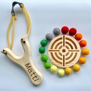 May include: A wooden slingshot with the name "Matti" engraved on it, next to a wooden target with a bullseye design surrounded by colourful felt balls.