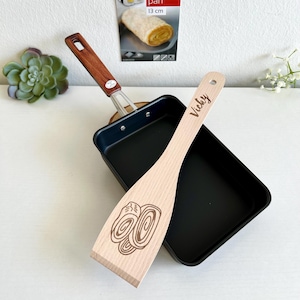 Può includere: Padella tamago nera, una padella giapponese per omelette, con una spatola di legno incisa con la parola "Vicky". La padella ha un diametro di 13 cm. La spatola ha un disegno di un'omelette arrotolata.