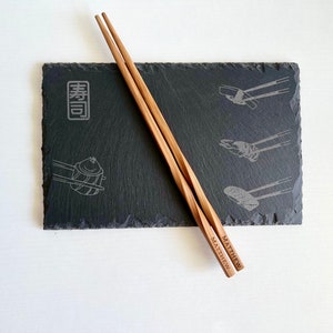 Puede incluir: Un par de palillos de madera con el nombre "Matthew" grabado en ellos. Los palillos están apoyados en una bandeja de servir de pizarra negra con un diseño de sushi y la palabra "粋" en japonés.