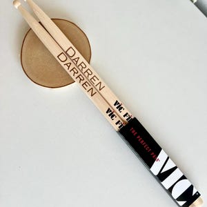 May include: A pair of wooden drumsticks with the names "Darren" and "Vic Firth" printed on them. The drumsticks are packaged in a black box with the text "Vic Firth The Perfect Pair".