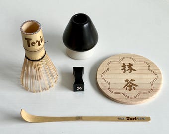 May include: A set of matcha tea ceremony tools including a bamboo whisk, a black ceramic bowl, a wooden stand with the text "抹茶" (Matcha) and "茶" (Tea) in Japanese, a black wooden whisk holder, and a bamboo matcha scoop with the text "Tori" engraved on it.