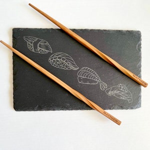 Könnte beinhalten: Ein schwarzes Sushi-Servierbrett aus Schiefer mit gravierten Sushi-Illustrationen und zwei Essstäbchen aus Holz. Die Essstäbchen sind hellbraun mit einem gedrehten Design und dem eingravierten Namen "MATTHEW".