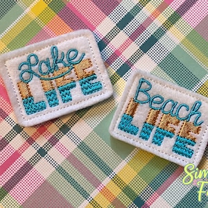 May include: Two white felt patches with embroidered text. The top patch says "Lake Life" in gold and blue lettering. The bottom patch says "Beach Life" in gold and blue lettering. The patches are on a plaid background.