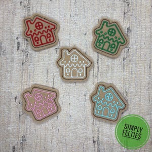 May include: Five felt gingerbread house cutouts arranged in a cluster. Each tan felt shape features embroidered outlines in red, green, white, pink, and light blue, depicting roof details, windows, and doors on each individual house shape.