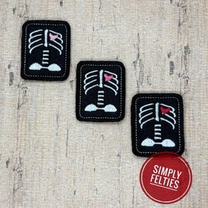 May include: Three rectangular black felt patches featuring white embroidered rib cage designs. Each patch displays a small heart in a different shade of pink or red positioned within the rib cage. A red circular logo reads SIMPLY FELTIES.