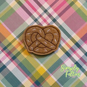 May include: A brown felt pretzel with white felt sprinkles. The pretzel is shaped like a heart and is on a plaid background.