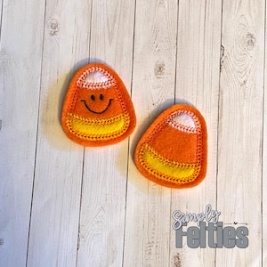 May include: Two felt candy corn ornaments, one with a smiling face, on a white wood background. The text "Simply Felties" is visible in the bottom right corner of the image.