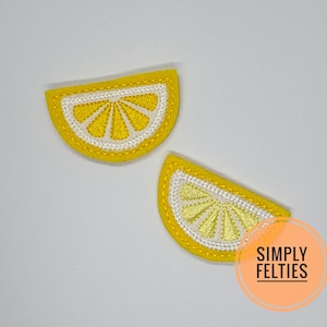 May include: Two embroidered lemon slice patches. Each patch features a yellow rind, a white inner section, and a yellow center with a stitched design. The patches are on a white background. The patches are from Simply Felties.