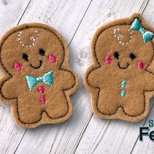 May include: Two brown felt gingerbread man ornaments with black stitched outlines. One has a blue bow tie and pink cheeks, the other has a blue bow and blue teardrops. Both have stitched smiles and white stitched details.