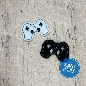 May include: Two felt patches shaped like video game controllers, one white with black stitching and details, and one black with white stitching and details. The patches feature embroidered directional pads and buttons. A blue circular logo reads SIMPLY FELTIES.