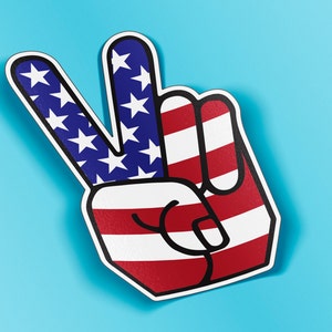 May include: A foam hand in the shape of the peace sign, featuring the American flag design. The fingers are blue with white stars, and the palm has red and white stripes. The hand is outlined in black and set against a light blue background.