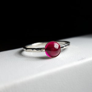 Puede incluir: Un anillo de plata con una banda martillada y una gema grande, redonda y de color rojo intenso. La gema es el punto focal, captando la luz y proporcionando un toque de color sobre la plata. El anillo es simple pero elegante.