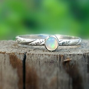 May include: A silver ring with an oval opal gemstone. The ring band has a delicate floral pattern. The opal has a milky white base with flashes of color. The ring is displayed on a wooden surface.
