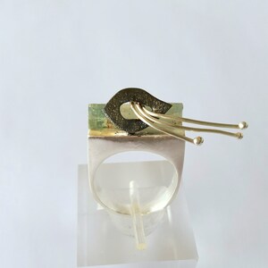 May include: A unique silver ring with an abstract design. The ring features a rectangular green stone with a dark, textured element and gold-colored accents. Three gold-colored wires extend from the top, each ending in a small sphere.