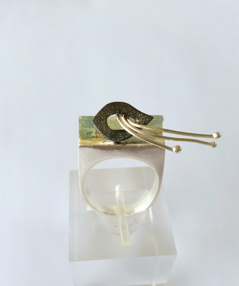 May include: A unique silver ring with an abstract design. The ring features a rectangular green stone with a dark, textured element and gold-colored accents. Three gold-colored wires extend from the top, each ending in a small sphere.
