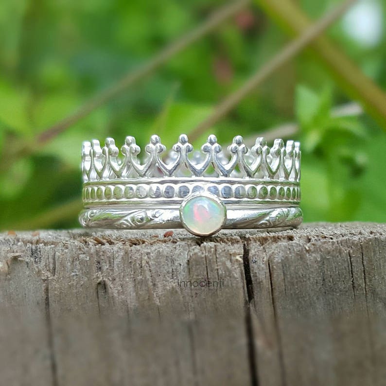May include: A stack of three silver rings. The top ring is shaped like a crown. The middle ring has an opal gemstone. The bottom ring has a floral design. The rings are on a wooden surface with a green background.