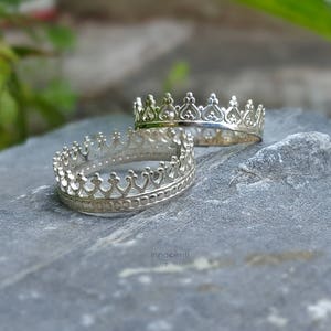 May include: Two silver crown rings sit on a grey stone surface. The rings feature a detailed design with small, pointed arches and decorative elements. The background shows blurred green foliage and hints of pink flowers.