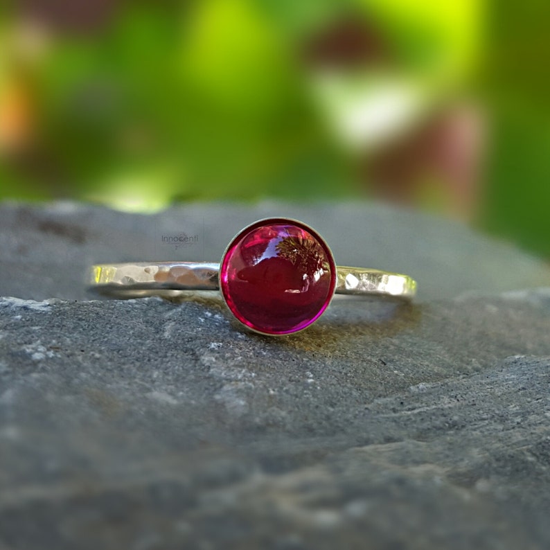 Puede incluir: Un anillo de plata con una banda martillada y una gran gema redonda de color rojo intenso. La gema es el punto focal, reflejando la luz, sobre un fondo verde borroso. El anillo se muestra sobre una superficie de piedra gris.