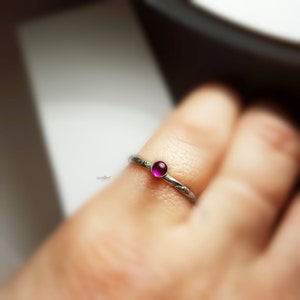 May include: A silver ring featuring a small, round, magenta gemstone. The ring's band has a delicate etched pattern. The ring is displayed on a finger, with a blurred background.