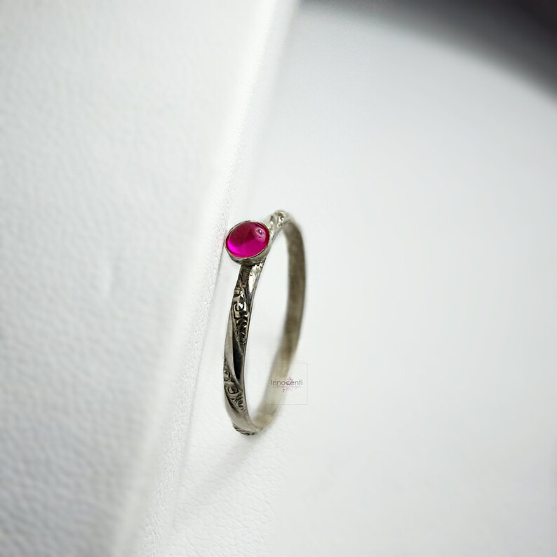 May include: A silver ring with a small, round, magenta gemstone. The band has an etched floral pattern. The ring is set against a white backdrop.