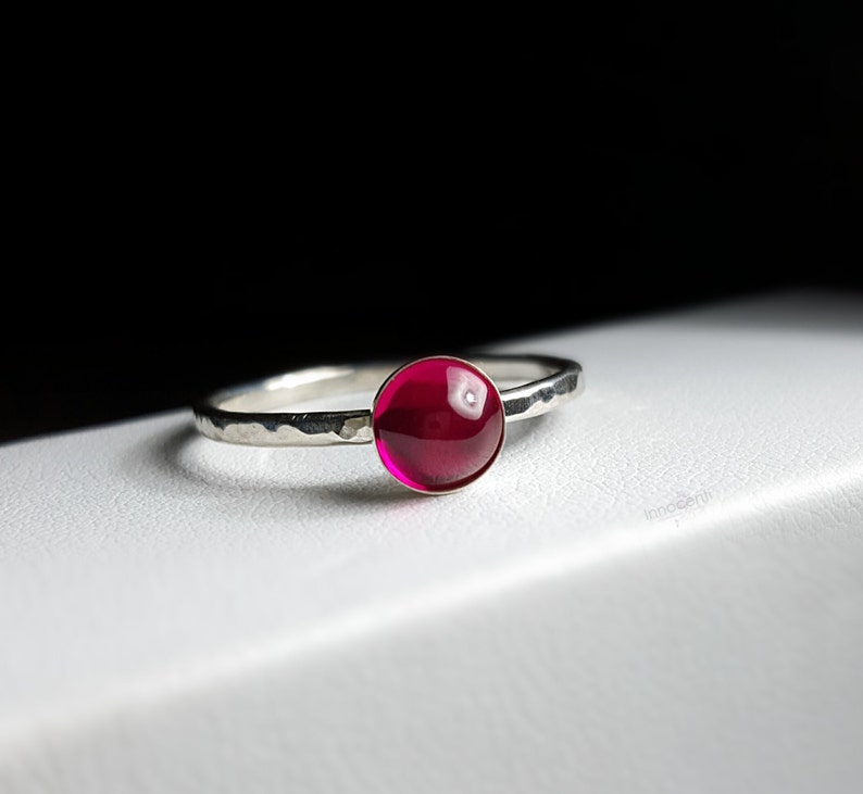 Puede incluir: Un anillo de plata con una banda martillada y una gema grande, redonda y de color rojo intenso. La gema es el punto focal, captando la luz y proporcionando un toque de color sobre la plata. El anillo es simple pero elegante.