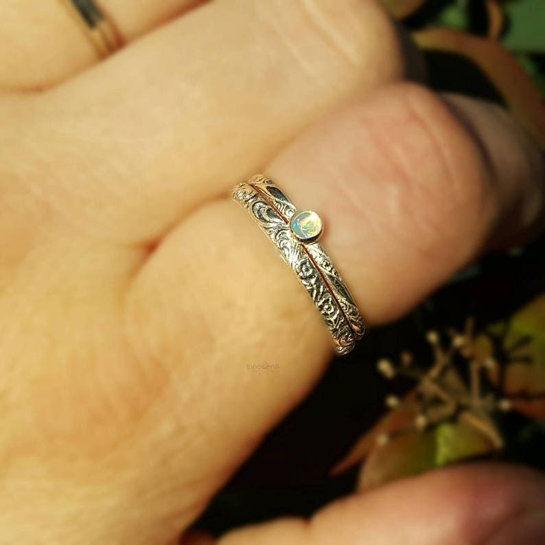 May include: A silver ring with an ornate, engraved design. The ring features a small, round, light-colored gemstone. The ring is on a finger, with a blurred background of greenery.