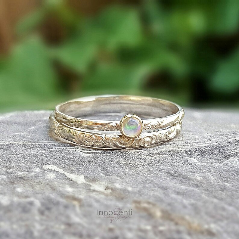 May include: Two silver rings, one with a small, round opal gemstone. The rings are stacked, with the bottom ring featuring an engraved floral pattern. The opal has a subtle play of color. The rings are set against a blurred green background.