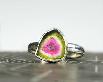 Könnte beinhalten: Ein Silberring mit einem Wassermelonen-Turmalin-Edelstein. Der Edelstein hat einen rosa Kern, einen grünen äußeren Rand und ein weißes Band dazwischen. Der Ring ist in einem polierten Silberband gefasst.