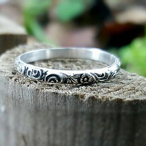 May include: A silver ring with an engraved floral pattern. The ring is set on a wooden surface, with a blurred green background. The ring's design features detailed swirls and flower motifs, showcasing a vintage or antique style.
