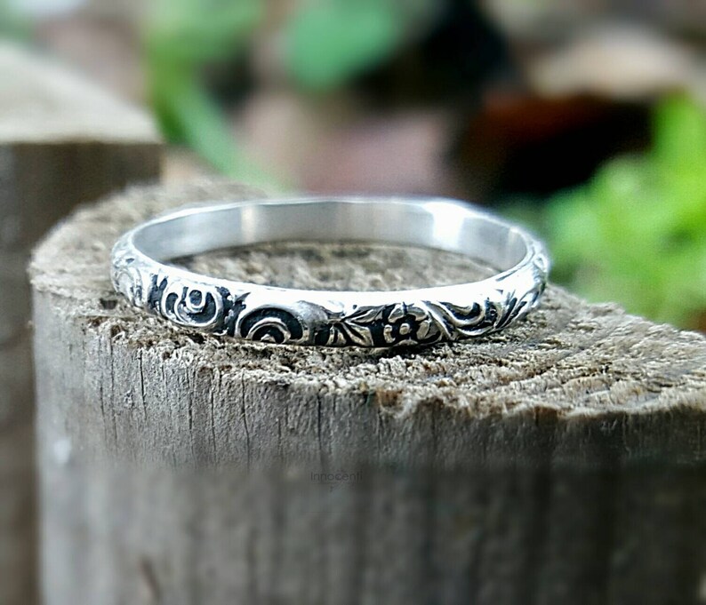 May include: A silver ring with an engraved floral pattern. The ring is set on a wooden surface, with a blurred green background. The ring's design features detailed swirls and flower motifs, showcasing a vintage or antique style.